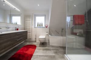 Modernes badezimmer dach mit Doppelwaschbecken, wandmontierter Toilette, Badewanne, Glasdusche, Bodenbelag in Holzoptik und auffälligen roten Akzenten wie Teppich und Handtuch.