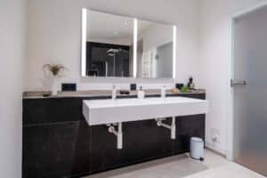 Ein modernes Badezimmer mit Doppelwaschbecken, großem beleuchtetem Spiegel, schwarz-weißem Dekor, Glasdusche, Topfpflanzen und minimalen Accessoires - perfekt, um Ihren Individualismus im Alltag zum Ausdruck zu bringen.