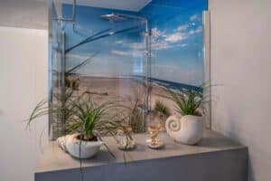 Der moderne Duschbereich ist mit einem Wandgemälde mit einer Strandszene ausgestattet, das Urlaubsfeeling pur vermittelt, und auf einem nahe gelegenen Sims stehen Topfpflanzen und dekorative Muscheln.