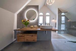 Modernes Bad mit rundem, hinterleuchtetem Spiegel, hölzernem Waschtisch, Gefäßwaschbecken, Tulpenvase und begehbarer Glasdusche unter einer schrägen Decke - perfekt für Wohnungen in Hanglage.