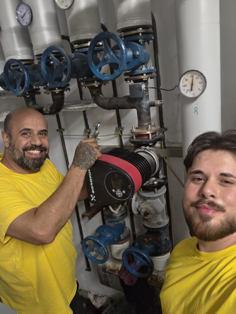 Zwei Männer in gelben Hemden stehen vor Industrierohren und Ventilen; einer hält ein Durchflussmessgerät in der Hand. Beide lächeln in die Kamera, was den Teamgeist hinter unserer "Über uns"-Geschichte widerspiegelt.