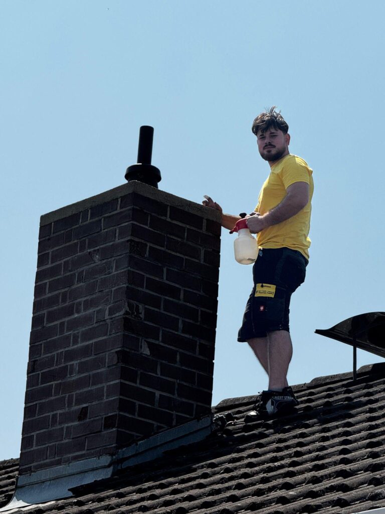 Eine Person in einem gelben Hemd steht auf einem Ziegeldach neben einem gemauerten Schornstein, hält eine Sprühflasche in der Hand und trägt Arbeitshandschuhe - und demonstriert damit das praktische Fachwissen, das hinter unserer über uns-Geschichte steckt.