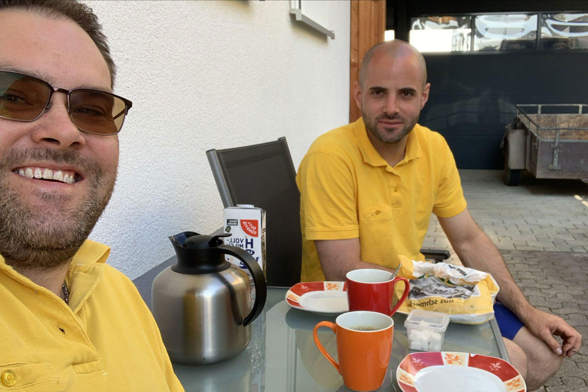 Zwei Männer in gelben Hemden sitzen an einem Tisch im Freien mit Kaffeebechern, einer Kaffeekanne und Essen und lächeln in die Kamera - vielleicht besprechen sie beim Frühstück ihr jüngstes Projekt zur Badsanierung.