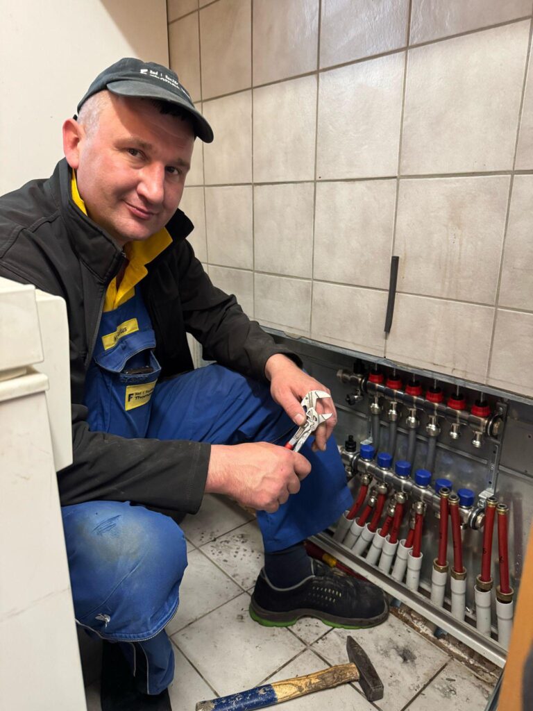 Ein Mann in Arbeitskleidung kniet auf einem gefliesten Boden und benutzt einen Schraubenschlüssel, um die Ventile eines Rohrverteilers in einem teilweise geöffneten Schrank einzustellen - ein Beweis für die Kompetenz von über uns.