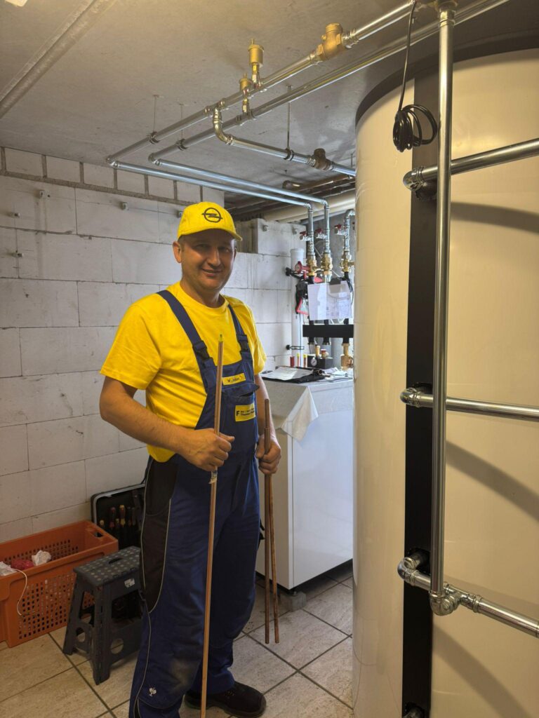 Ein Mann in einem gelben Hemd, einer Mütze und einem blauen Overall steht in einem Hauswirtschaftsraum und hält Rohre neben einem großen zylindrischen Tank und Sanitärarmaturen, bereit zu helfen.