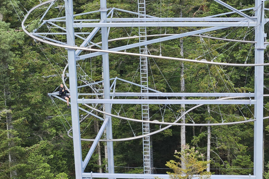 Ein metallener Aussichtsturm mit Wendeltreppe steht inmitten dichter grüner Bäume. Eine Person sitzt auf der Struktur in der Nähe der Treppe, vielleicht macht sie eine Pause von ihrer Arbeit und genießt die ruhige Aussicht.