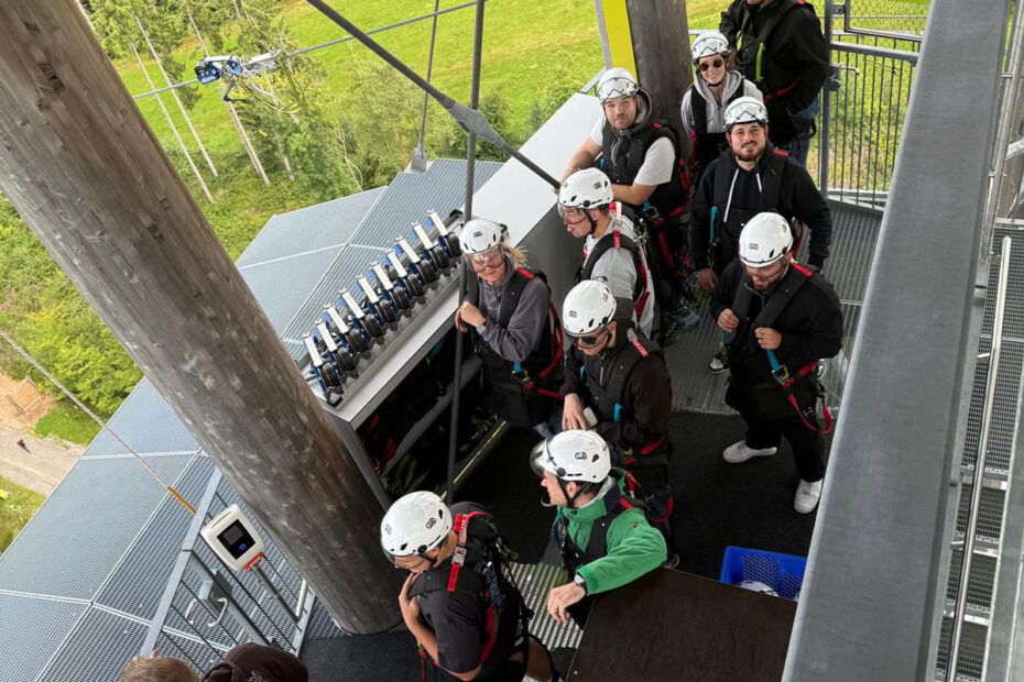 Eine Gruppe von Personen mit Helmen und Klettergurten wartet auf einer Metallplattform auf eine Outdoor-Abenteueraktivität, möglicherweise im Rahmen einer teambildenden Veranstaltung, umgeben von hohen Holzpfählen und Bäumen im Hintergrund.