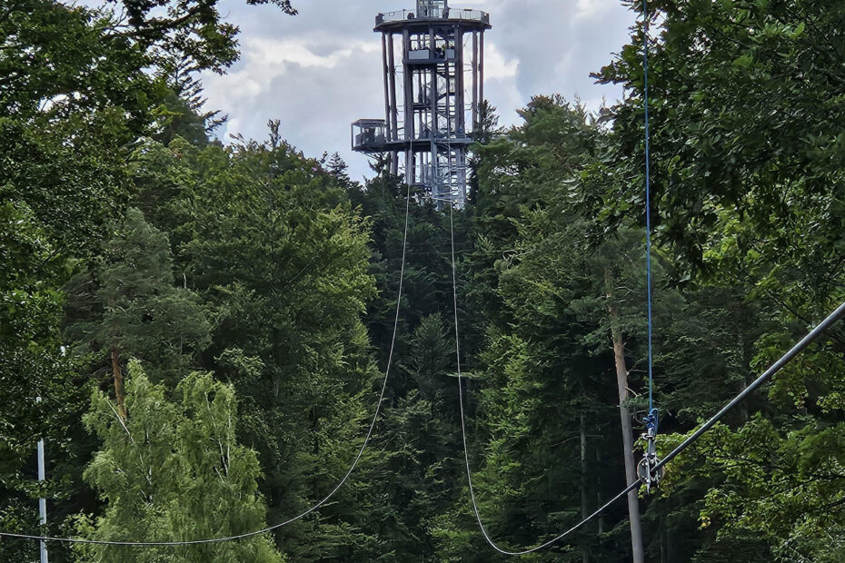 Ein hoher Aussichtsturm steht inmitten dichter grüner Bäume, und eine Seilrutsche führt durch den Wald auf den Turm zu - perfekt für alle, die den Nervenkitzel suchen oder einen abenteuerlichen Job in der Freizeit suchen.