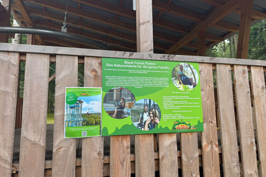 An einem Holzzaun ist ein Informationsschild für die Black Forest Flyline angebracht, auf dem Text und Bilder über die Attraktion und ihre unterhaltsamen, familienfreundlichen Aktivitäten zu lesen sind - perfekt für Ihr nächstes Abenteuer oder sogar einen Sommerjob.