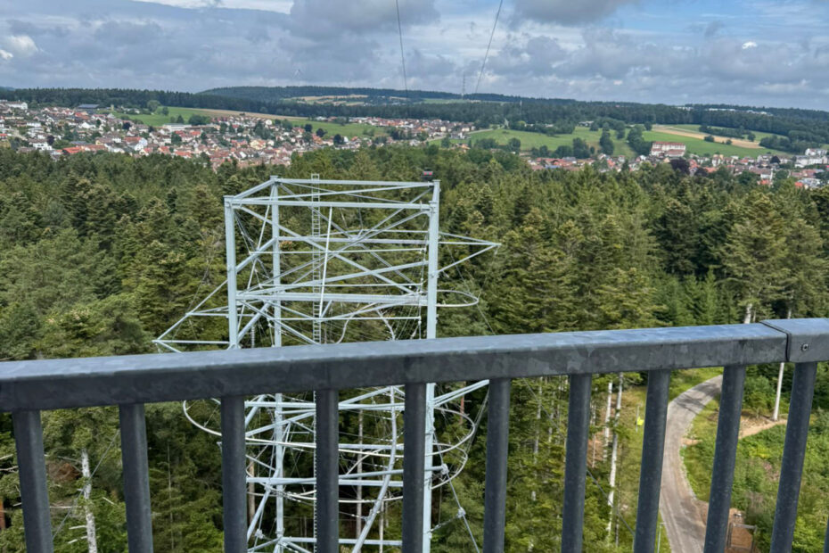 Blick von einem Metallgeländer auf einen spiralförmigen Aussichtsturm über einem Wald, mit einer Stadt und offenen Feldern in der Ferne unter einem teilweise bewölkten Himmel - der perfekte Ort, um nach einem langen Arbeitstag nachzudenken.