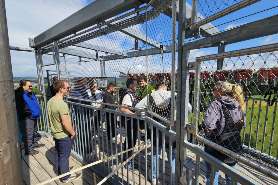 Eine Gruppe von Menschen steht auf einer umzäunten Aussichtsplattform im Freien und macht vielleicht eine Pause von ihrer Arbeit, während im Hintergrund Bäume und grüne Felder unter einem teilweise bewölkten Himmel zu sehen sind.